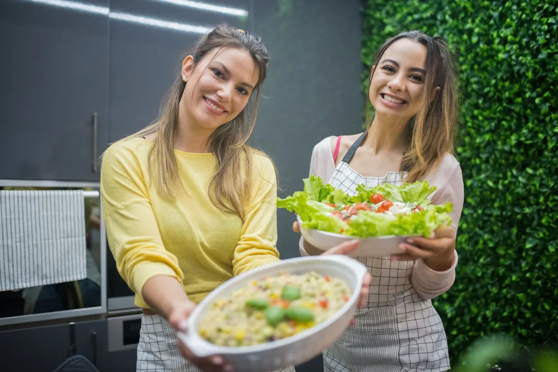 Two smiling women holding bowls of fresh vegetables and salad — healthy food choices that support gut health and mood